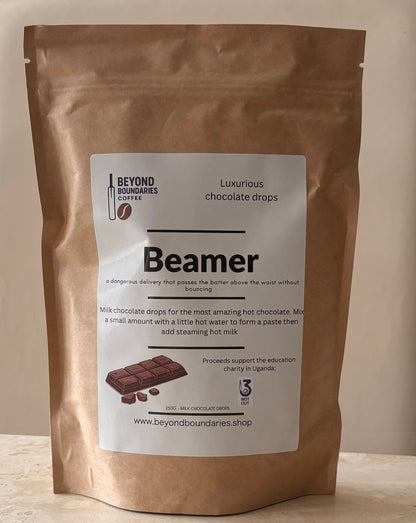 A stand-up kraft paper pouch of Beyond Boundaries Chocolate 'Beamer' chocolate drops, featuring a white label with product details and a chocolate bar illustration, set against a neutral beige background.