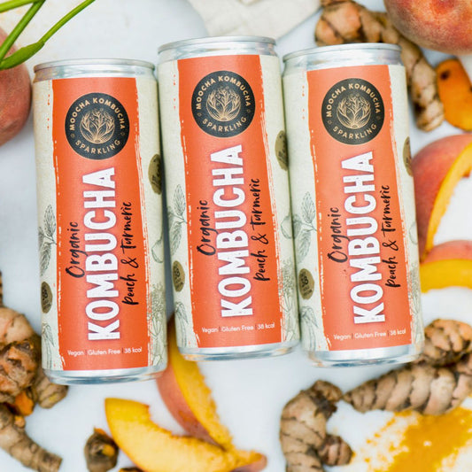 Three cans of Kombucha surrounded by peaches and turmeric on a white background