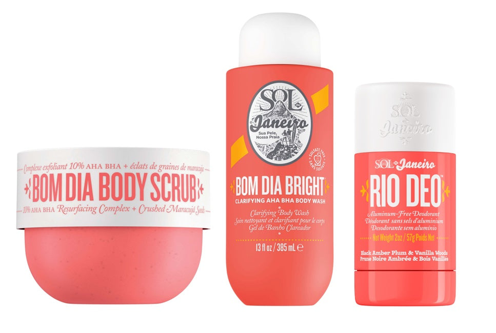 Three Sol Janeiro body care products on a white background: a round pink scrub container labeled 'BOM DIA BODY SCRUB,' a tall coral body wash bottle labeled 'BOM DIA BRIGHT,' and a matching coral deodorant stick labeled 'RIO DEO.'.